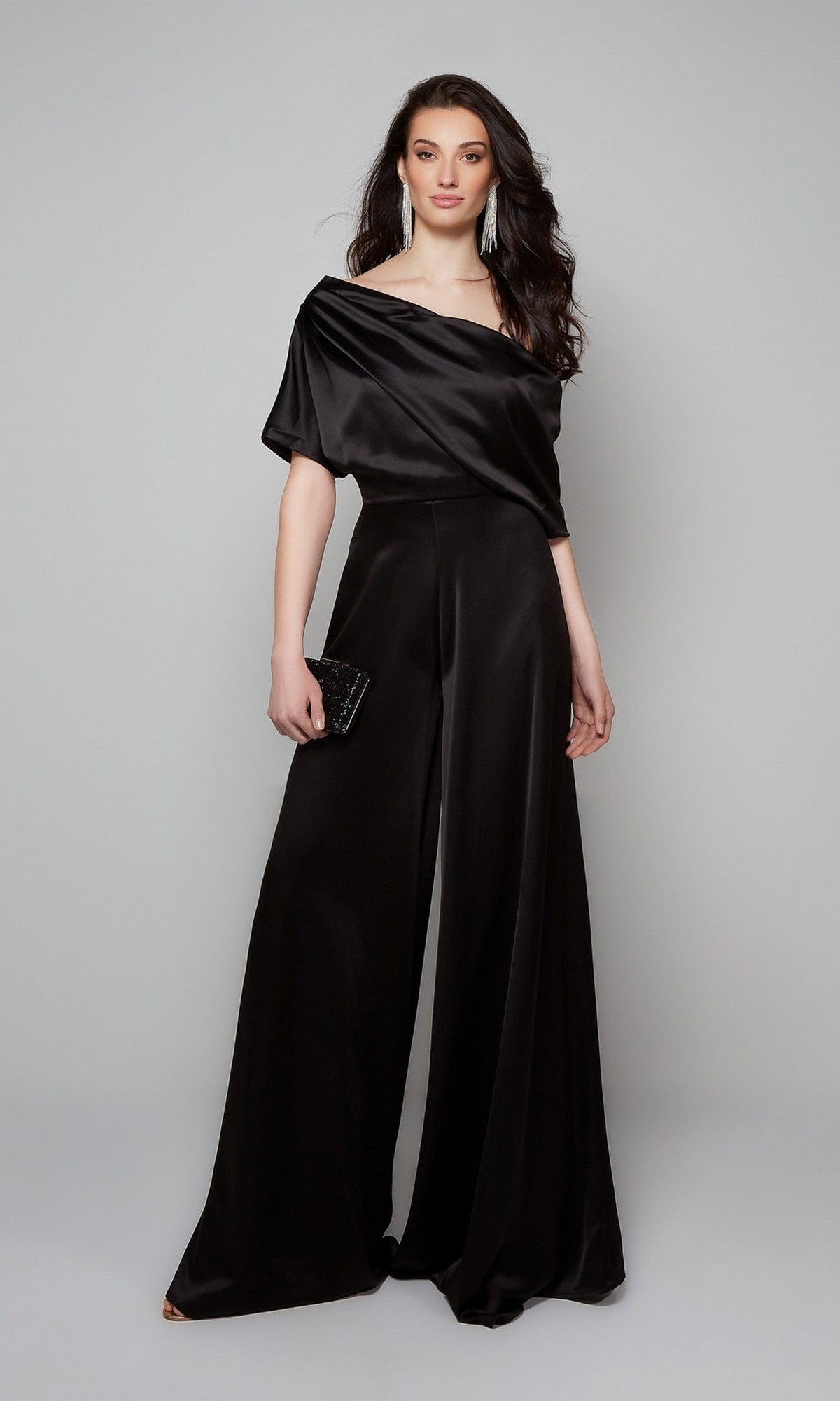 A woman stands against a plain background wearing the Alyce 70019 long formal jumpsuit with wide-leg pants, holding a black clutch and wearing long earrings.